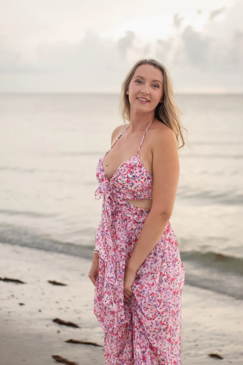 Woman in a pink floral dress smiling over her shoulder on the beach at golden hour