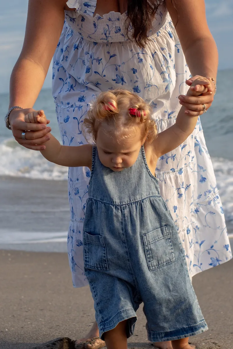 Toddler in denim overalls taking steps along the water while holding her mother's hands