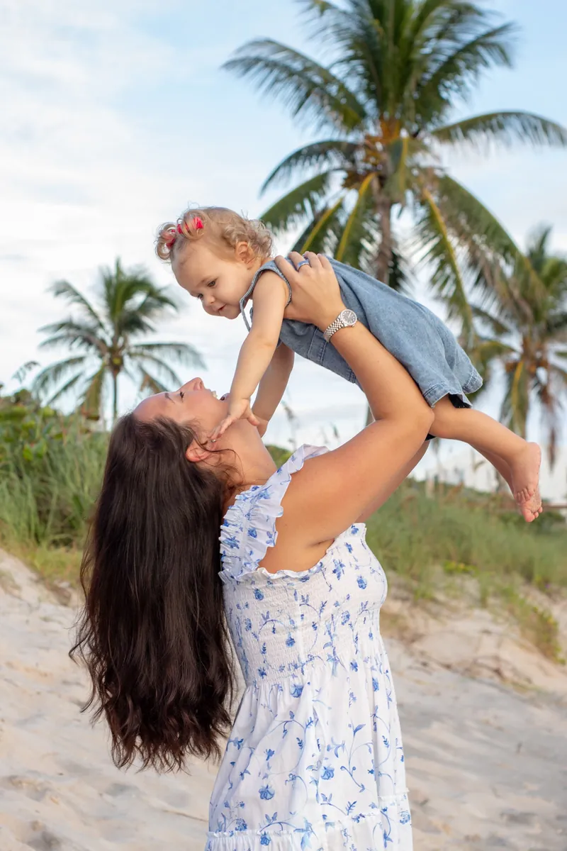Mother lifting her smiling toddler under palm trees during a family session in Jupiter, Florida